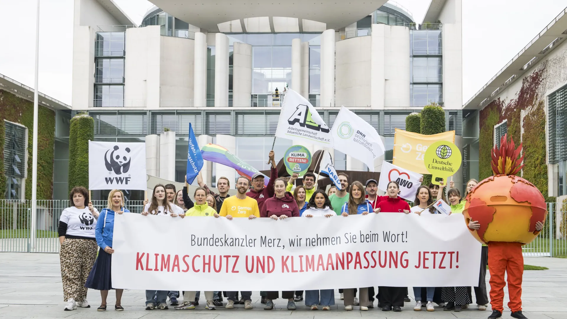 zwei Dutzend Umweltaktive vor dem Bundeskanzleramt in Berlin mit einem Banner, Aufschrift "Bundeskanzler Merz, wir nehmen Sie beim Wort! KLIMASCHUTZ UND KLIMAANPASSUNG JETZT!"; rechts im Bild, eine als brennende Erde verkleidete Person